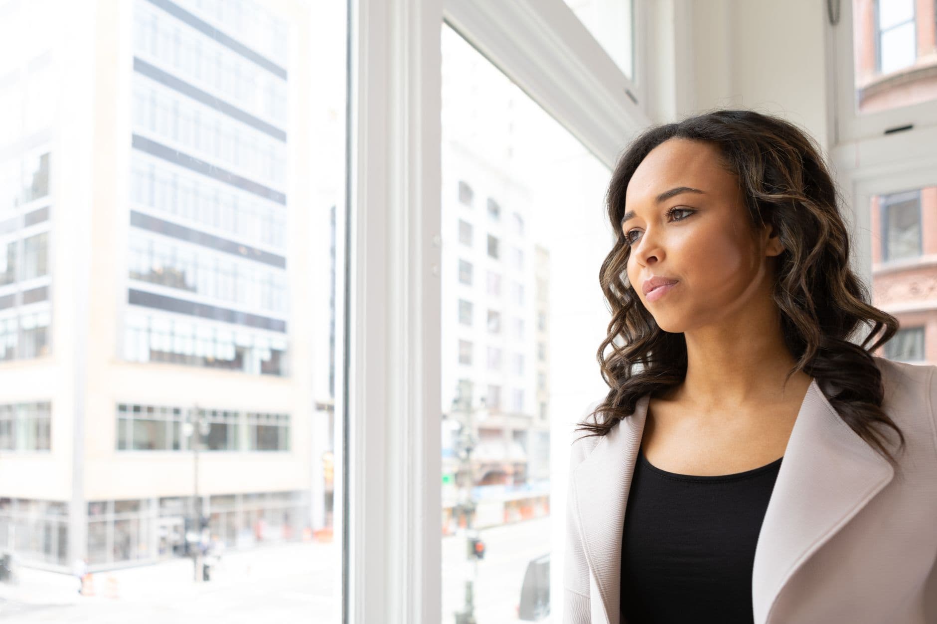 A professional woman gazing thoughtfully out an office window over the city, on mental health at work